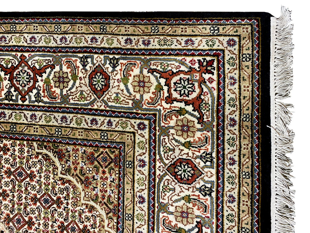 Detalle de la cenefa de una alfombra Indo-Tabriz marrón con ornamentos coloridos y flecos a la derecha