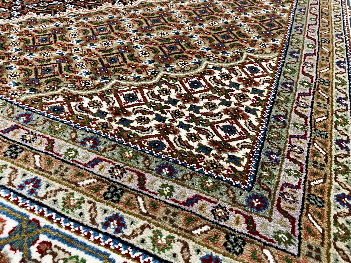 Esquina de una alfombra Indo-Tabriz marrón con adornos coloridos, cenefa decorativa y flecos.