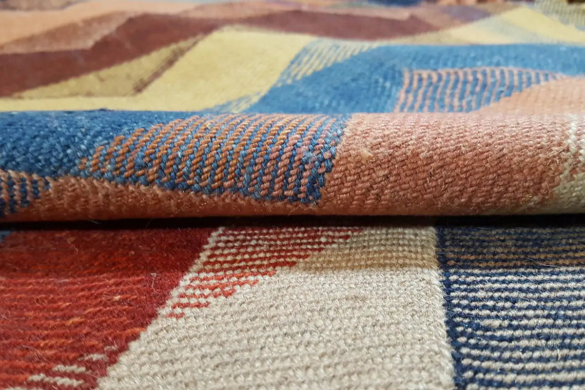 Primer plano: borde de alfombra con textura tejida y zigzag en azul, crema y rojo.