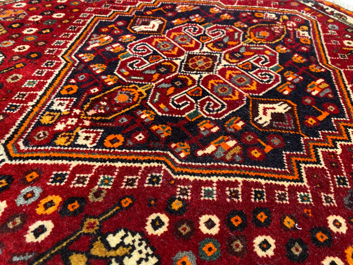 Alfombra Poshti de 60x60 cm, vista de cerca del ornamento central de colores sobre fondo rojo