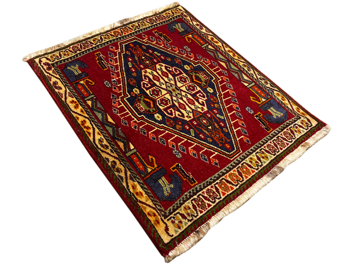 Alfombra Poshti (59×61 cm) extendida en diagonal, con patrón geométrico rojo y flecos