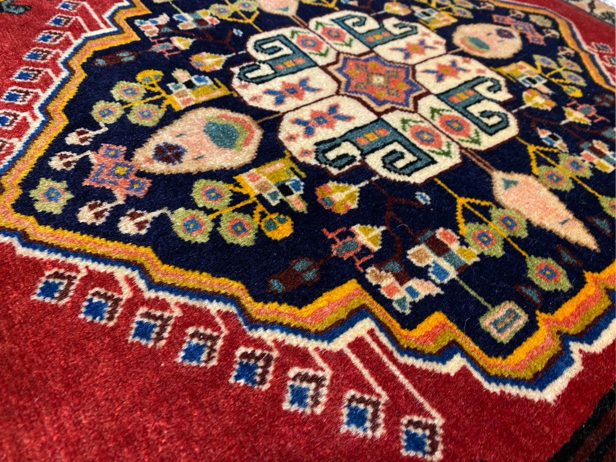 Alfombra Poshti con centro azul oscuro, bordes rojos y motivos geométricos coloridos