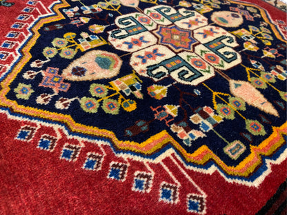 Alfombra Poshti con centro azul oscuro, bordes rojos y motivos geométricos coloridos