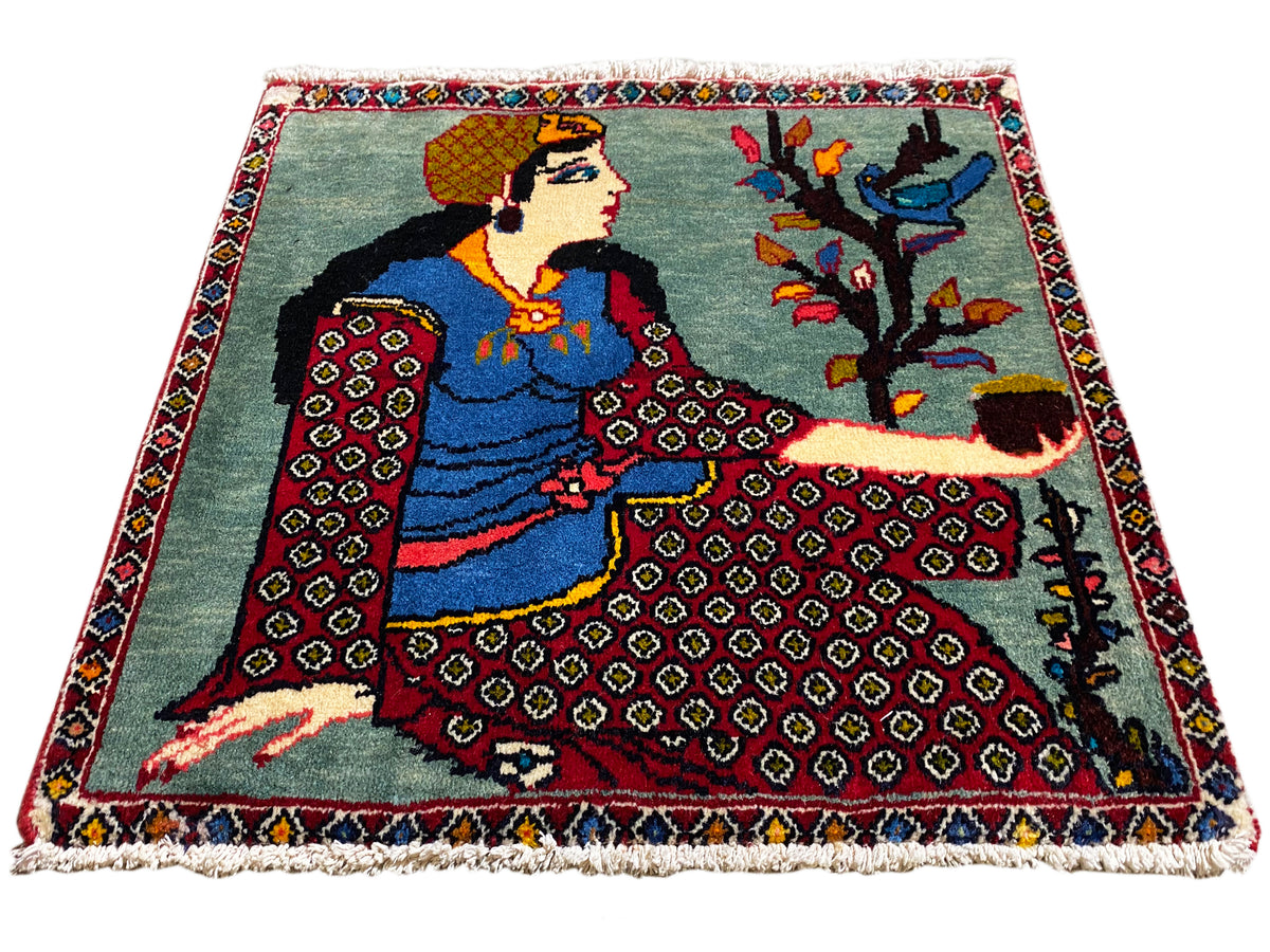 Alfombra Poshti de 68x63 cm en vista de tres cuartos con figura, árbol, bordes con motivos rojos y flecos