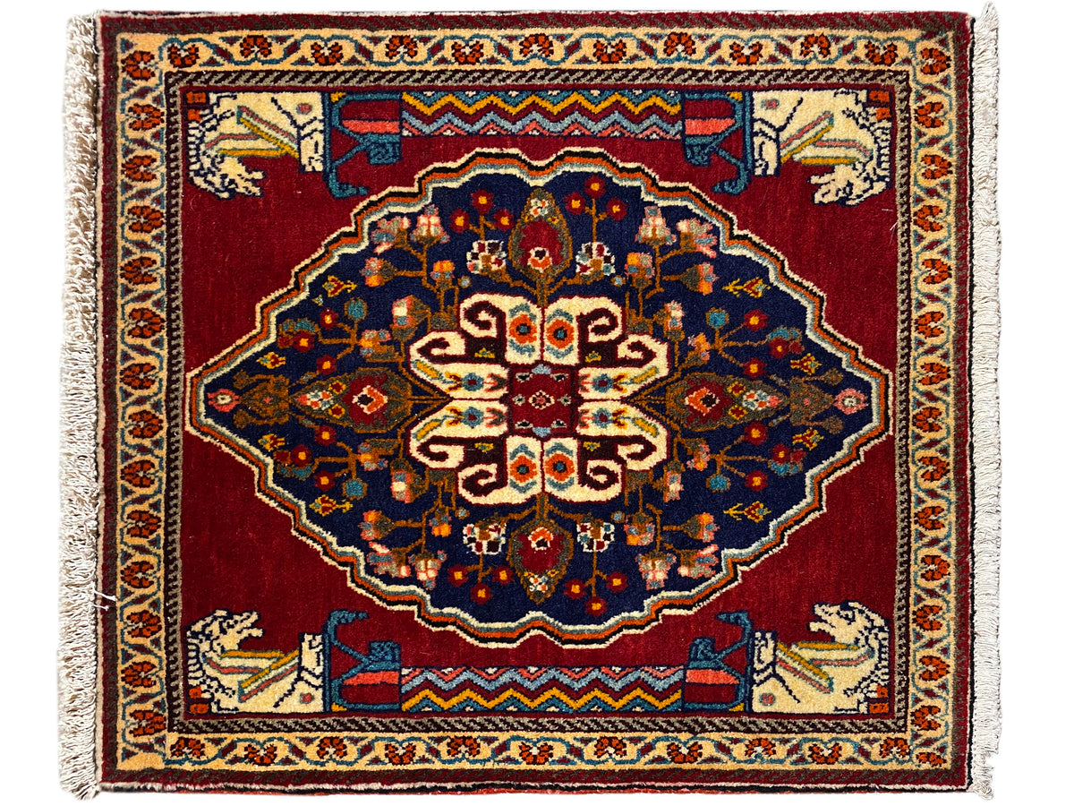 Alfombra rectangular con fondo rojo, motivos ornamentales azul oscuro y ribetes decorativos beige