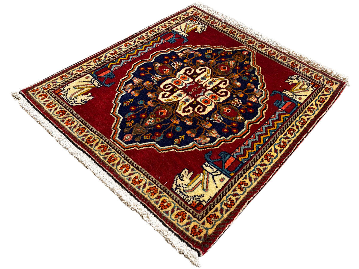 Alfombra con fondo rojo, motivo central azul oscuro, cenefas ornamentales beige y flecos