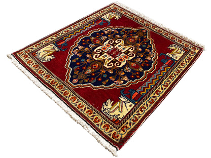 Alfombra con fondo rojo, motivo central azul oscuro, cenefas ornamentales beige y flecos