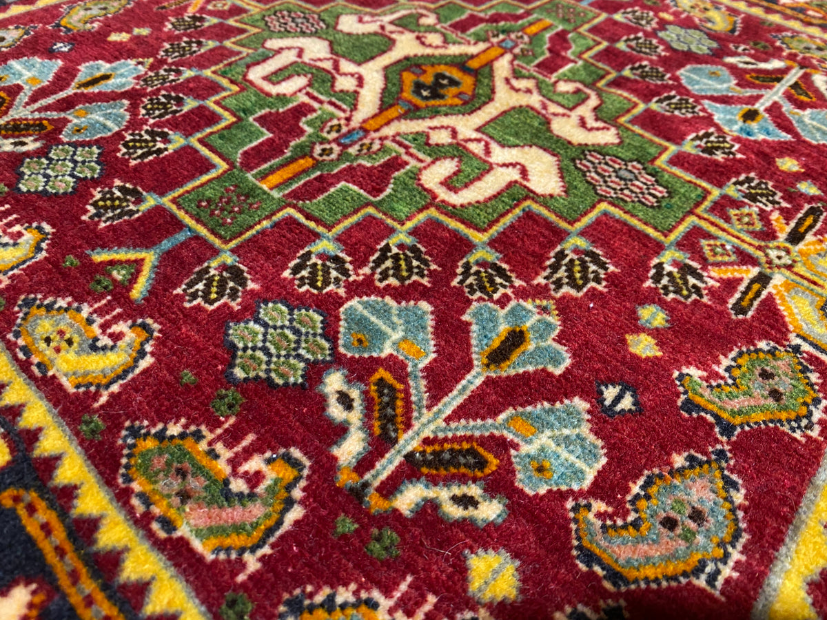 Alfombra Poshti de 61x61 cm en primer plano, campo rojo con motivos verdes y azules