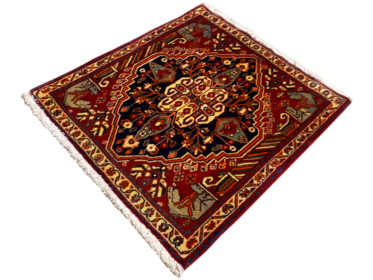Alfombra de 65x59 cm con fondo rojo, ornamentos centrales oscuros y flecos en los bordes