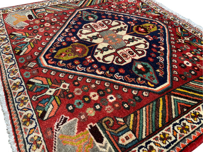 Alfombra oriental fotografiada en diagonal: diseño rojo, medallón oscuro y ribetes multicolores