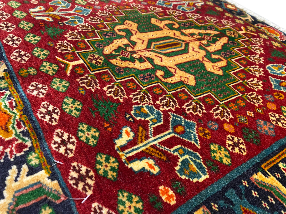 Detalle de la parte delantera de una alfombra con campo rojo, patrones en forma de estrella y líneas turquesas, pelo texturizado