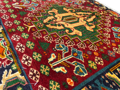 Detalle de la parte delantera de una alfombra con campo rojo, patrones en forma de estrella y líneas turquesas, pelo texturizado