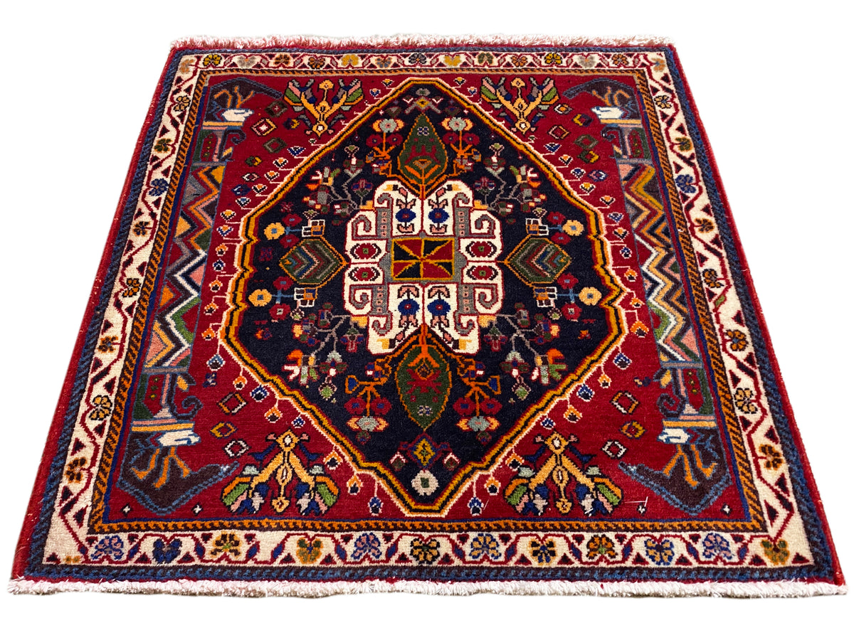 Alfombra Poshti (67×67 cm) en vista oblicua: cenefa, centro rojo, azul oscuro