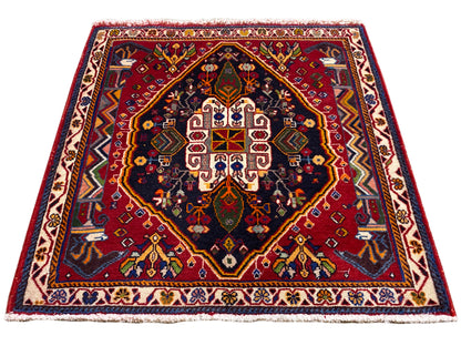 Alfombra Poshti (67×67 cm) en vista oblicua: cenefa, centro rojo, azul oscuro