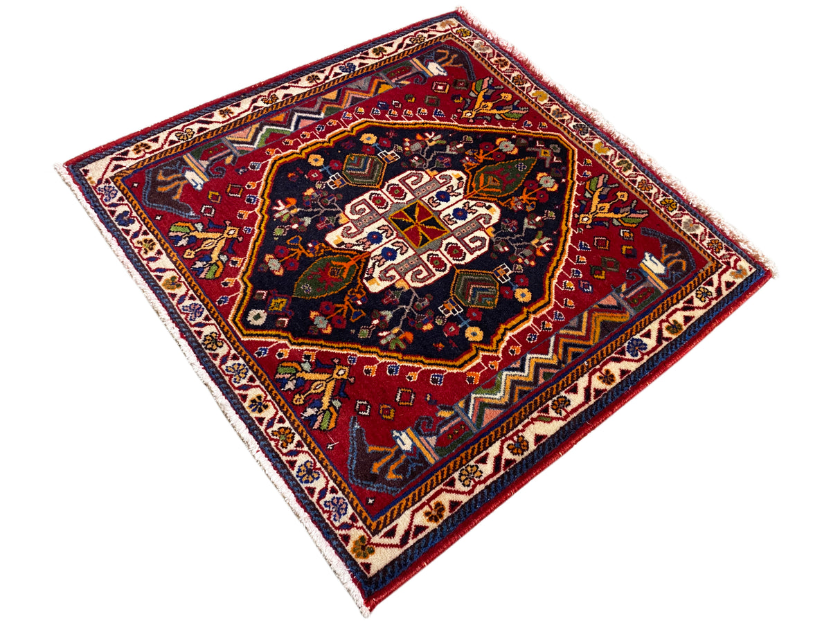 Alfombra Poshti (67×67 cm) en diagonal: fondo rojo, centro oscuro con motivos blancos