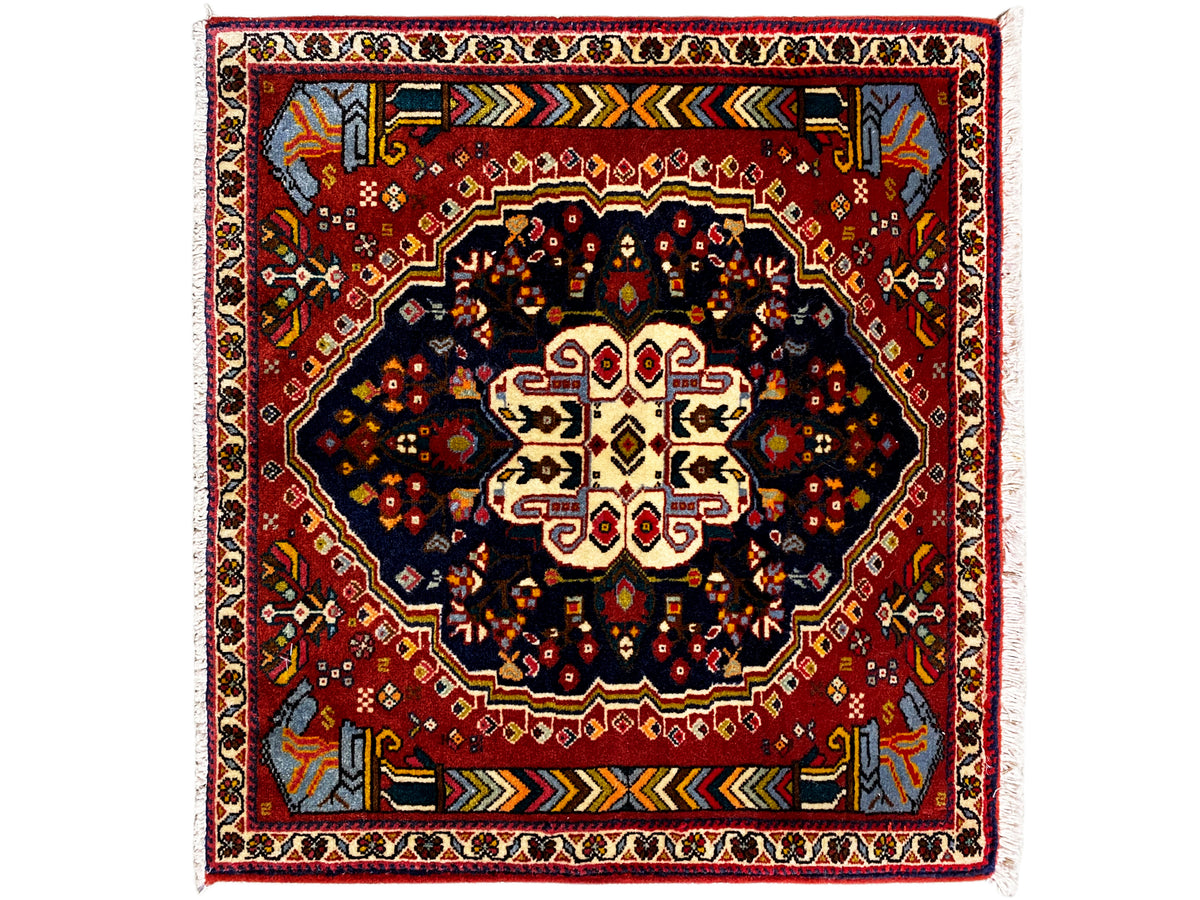Alfombra Poshti de 65x61 cm con borde rojo, motivo central oscuro y ornamentos coloridos