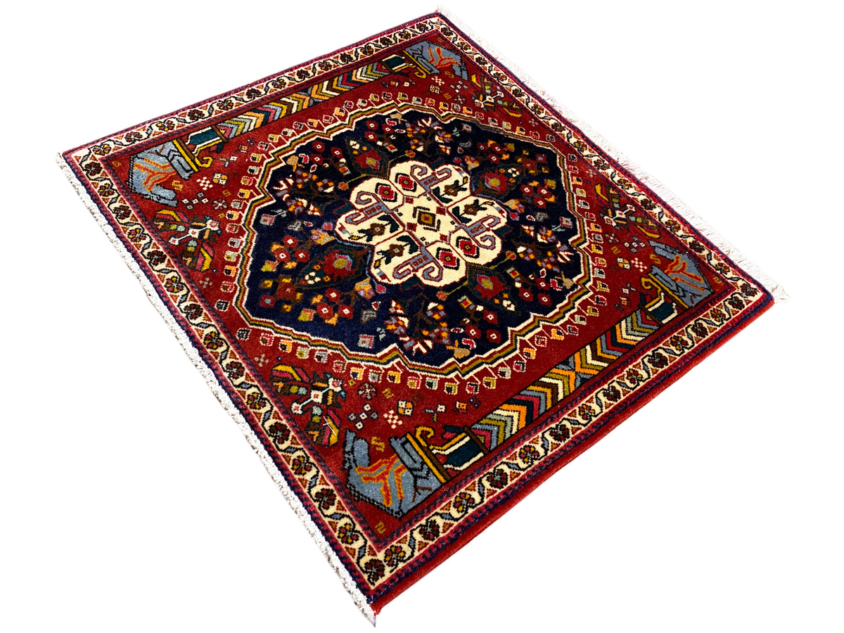 Alfombra Poshti de 65x61 cm, vista diagonal: campo rojo con un patrón oscuro y ornamentos en los bordes