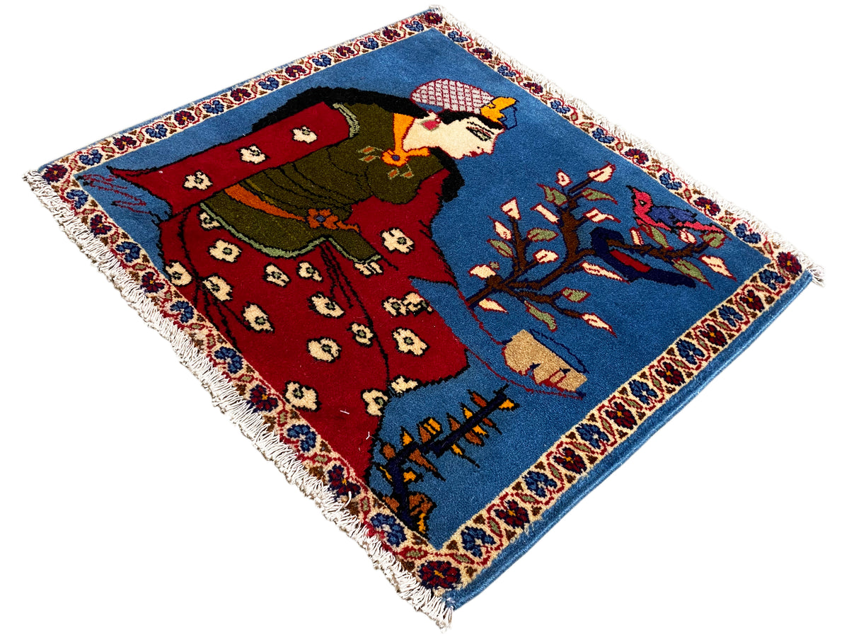 Alfombra Poshti de 65x63 cm vista en diagonal, figura, vestimenta roja y ornamentos azules